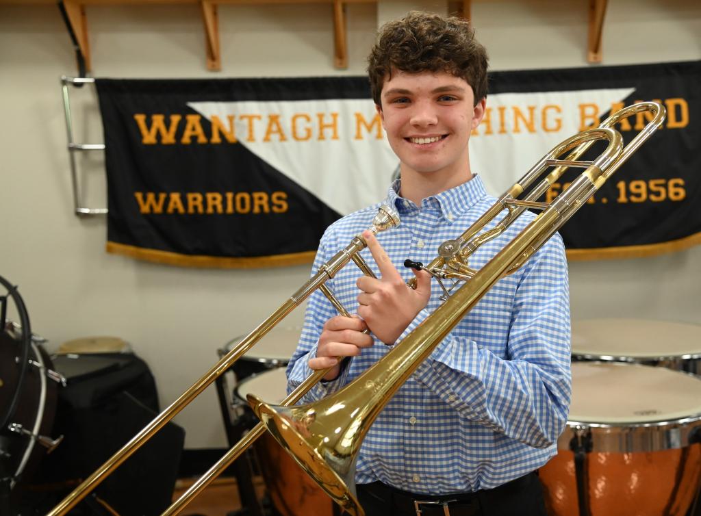 A smiling young man in a blue plaid shirt holds a brass trombone in a band room.